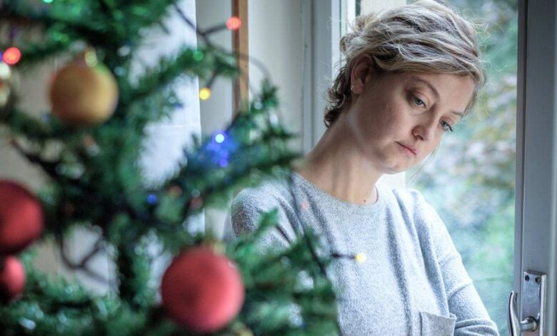 a young white woman looking sad and depressed leaning against a glass door with a decorated Christmas tree in the foreground x .jpg