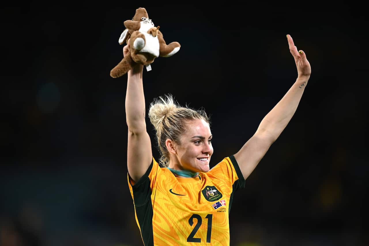 A woman in yellow football shirt holds both hands above her head.