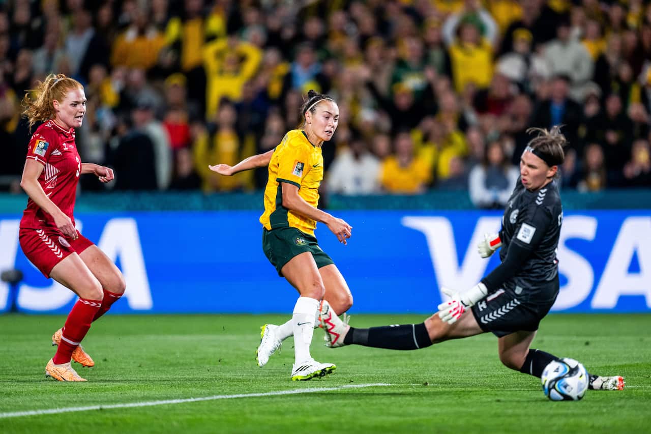 A woman in a yellow football uniform kicks a ball between the legs of a woman in a black football uniform