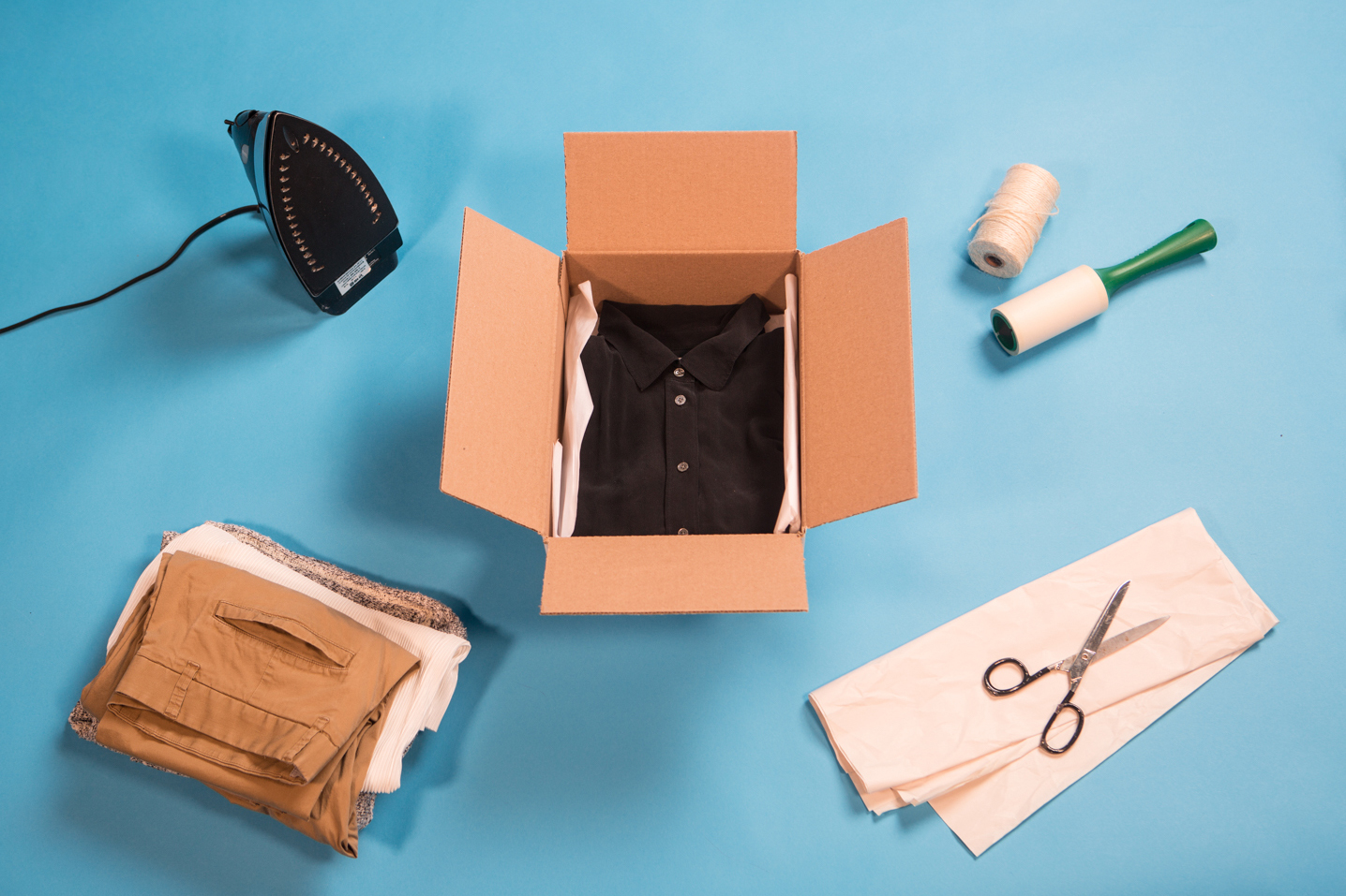 A black collared shirt is neatly folded inside a cardboard box on a blue background in the middle of the frame. The box is surrounded by an iron, a stack of clothes, a pair of scissors, tissue paper, twine and a lint roller.