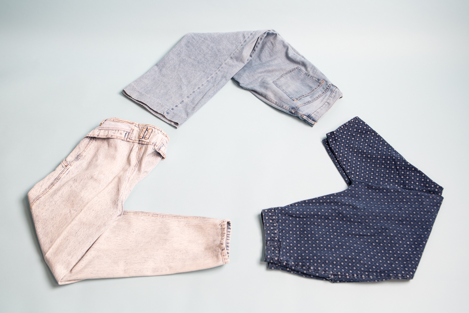 Three pairs of jeans in different shades of denim are arranged in the shape of the &quot;recycle&quot; logo on a light blue backdrop.